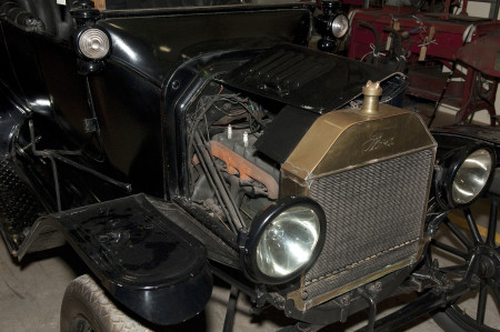A closeup of the engine of the car from the first image with the hood lifted. The front grill is inscribed with the brand name Ford. On either side are two round headlights. The interior of the engine has black wiring and an orange pipe or tube. 